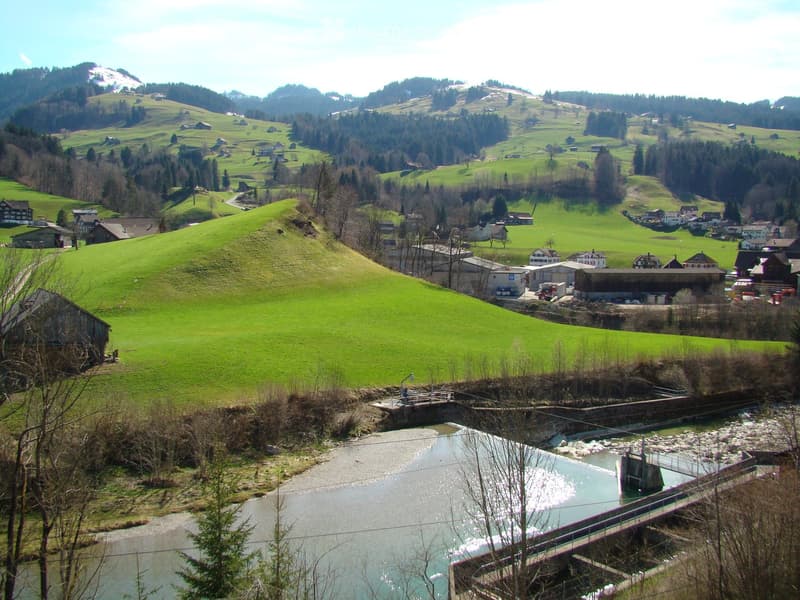 A scenic rural landscape with green hills, scattered houses, a river, and a bridge, surrounded by trees and mountains.