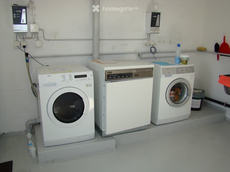 Three white washing machines and dishwasher, electrics and pipes, plastic waste bin, cleaning tools on wall