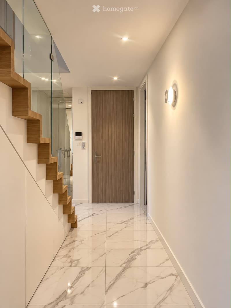Modern hallway with white marble flooring, wooden staircase with glass railing, and recessed lighting. Features a wooden door and wall-mounted light fixture.
