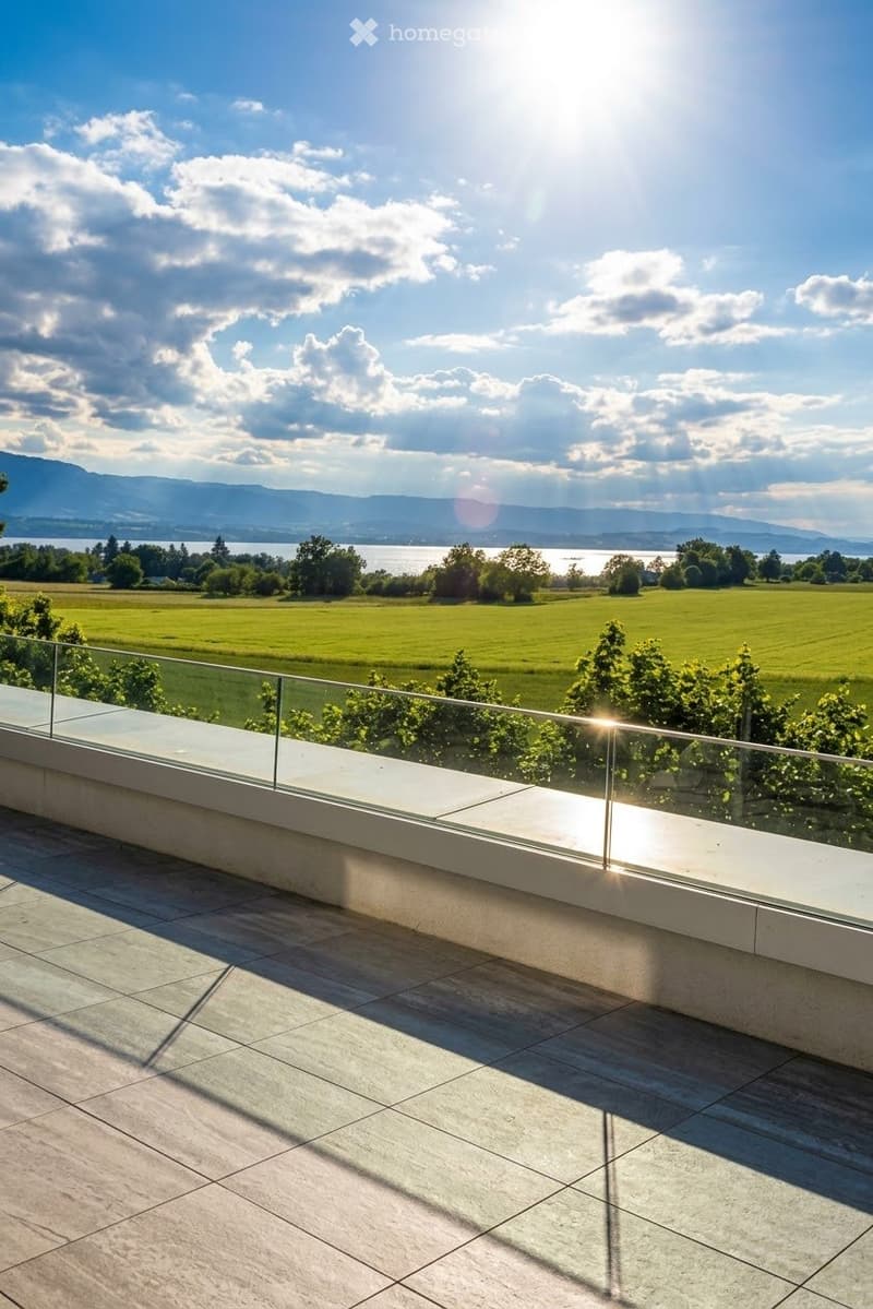 Modern balcony with glass railing, tiled floor, and scenic view of a green field, trees, and distant lake and mountains.