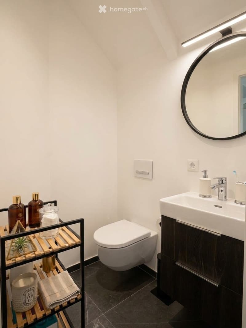 Modern bathroom with white walls, dark tiled floor, a white sink with a modern faucet, a toilet, a large round mirror, and a wooden shelf holding decorative items and toiletries.