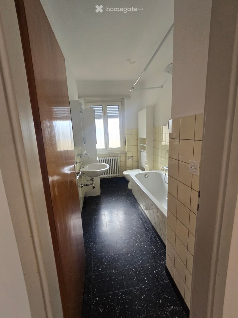Bathroom with black tiled floor, white bathtub, sink, toilet, and radiator. Light-colored tiles on walls and a window with blinds.