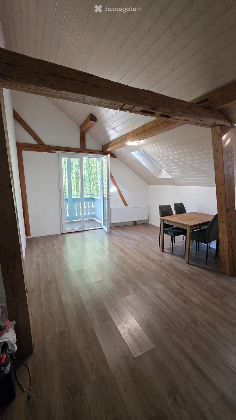Bright, airy attic space with wooden beams, wooden flooring, and a large skylight. A small dining area with a table and chairs is visible, and a sliding glass door opens to a balcony.