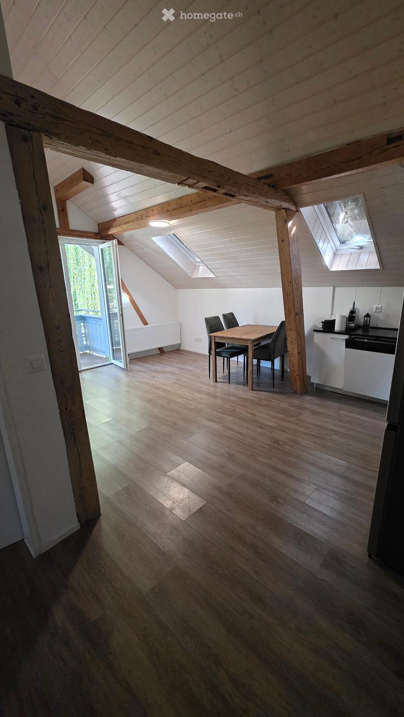 Bright, wooden-floored attic space with exposed wooden beams, skylights, a small dining area with a table and chairs, and a compact kitchen area with a counter and appliances.