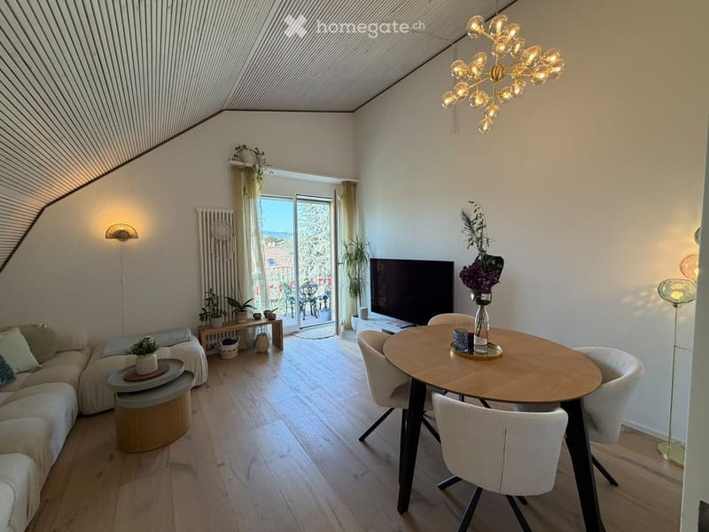 Modern living and dining area with a slanted ceiling, large windows opening to a balcony, a sectional sofa, a coffee table, a dining table with chairs, a TV, and decorative plants.