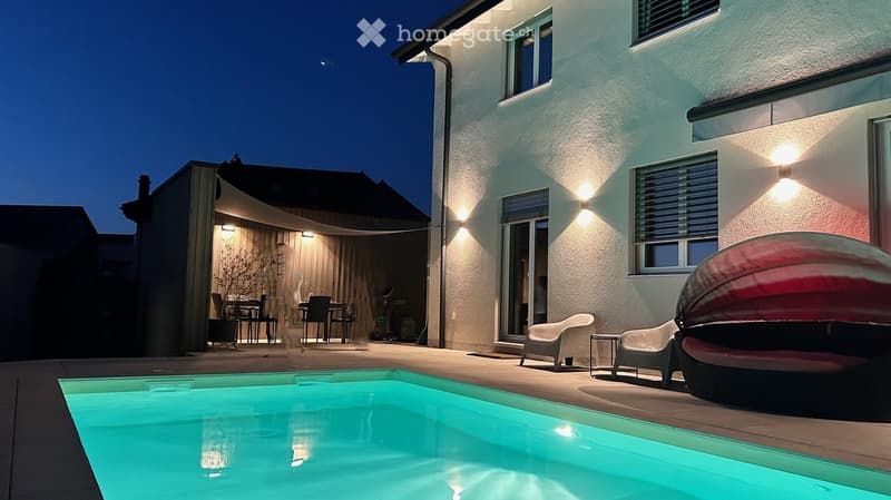 Evening view of a modern house with a swimming pool, outdoor seating area, and covered patio with dining furniture.