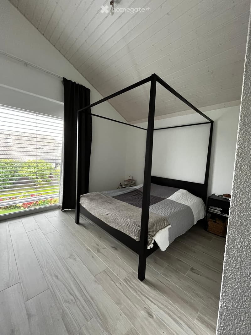 Modern bedroom with a raised platform bed, nightstand, and large window with blinds. The room features light-colored wooden flooring and white walls.
