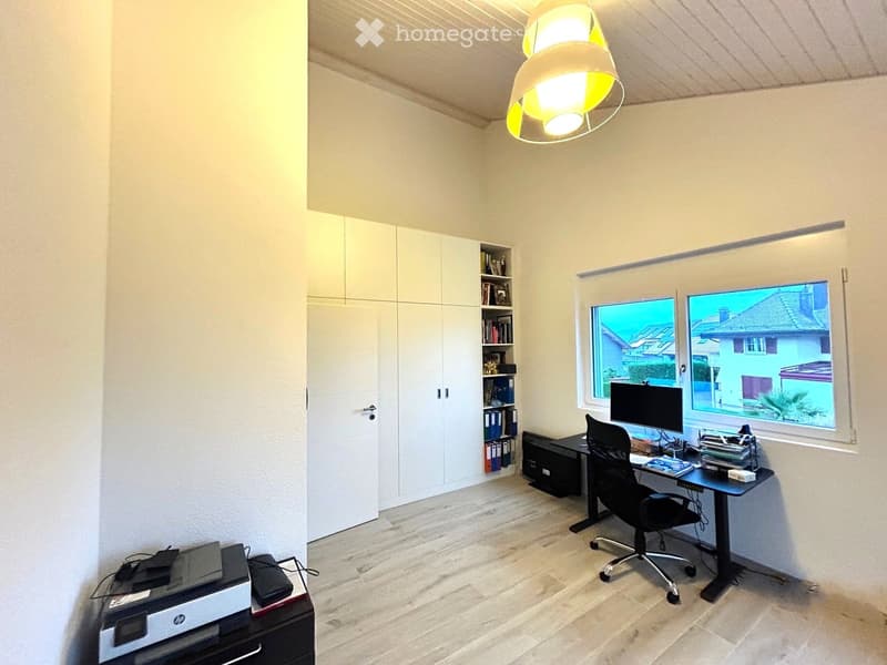 Modern office space with a desk, chair, computer, printer, and built-in cabinets and bookshelf. Large windows offer a view of the neighborhood.