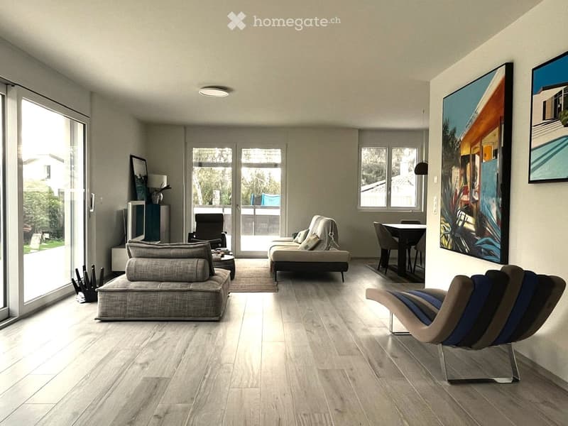 Modern living room with light-colored wood flooring, large windows, and a mix of seating including a sofa, chairs, and a dining table. Artwork adorns the walls, and the space is well-lit with natural light.