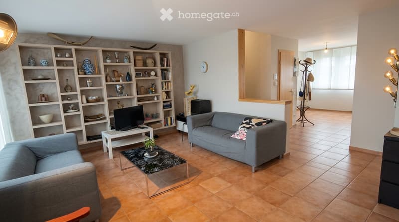 Living room with two sofas, coffee table, TV, bookshelf, and decorative items. The space features tiled flooring and large windows.