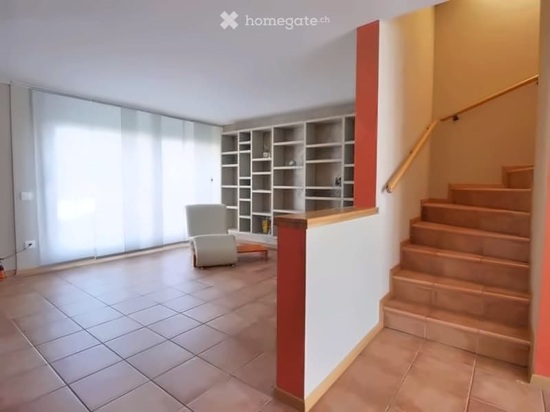 A modern, minimalist living room with a white sofa, a small table, and a built-in bookshelf. The room features tiled flooring and a staircase leading upwards.