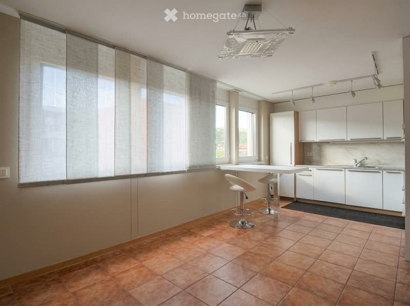 Modern kitchen with white cabinets, a central island, two stools, and a refrigerator. The space features large windows with blinds and a hanging light fixture.