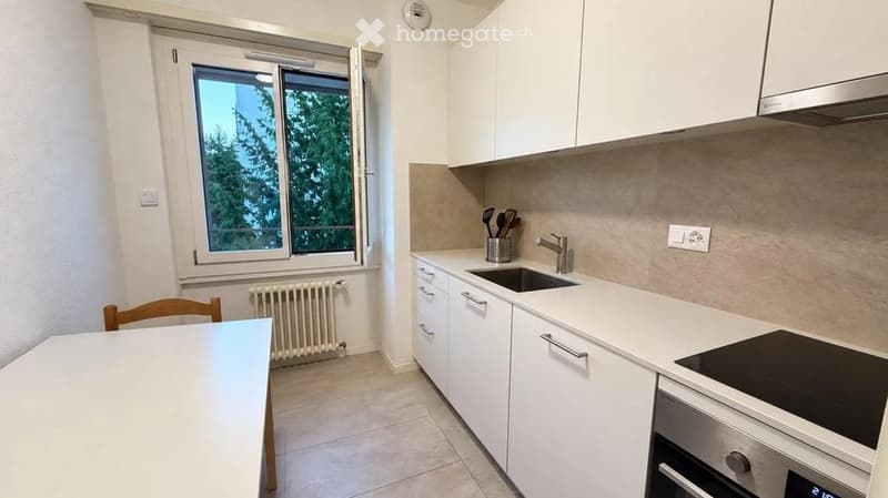 Modern kitchen with white cabinets, a sink, stove, oven, and a small dining area with a table and chair. The room features a large window with a view of greenery outside.