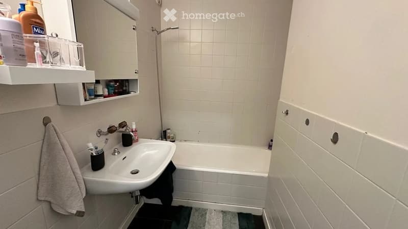 A small bathroom with a white tiled wall, a sink, a bathtub, a mirror, a cabinet, and a towel hanging on the wall.