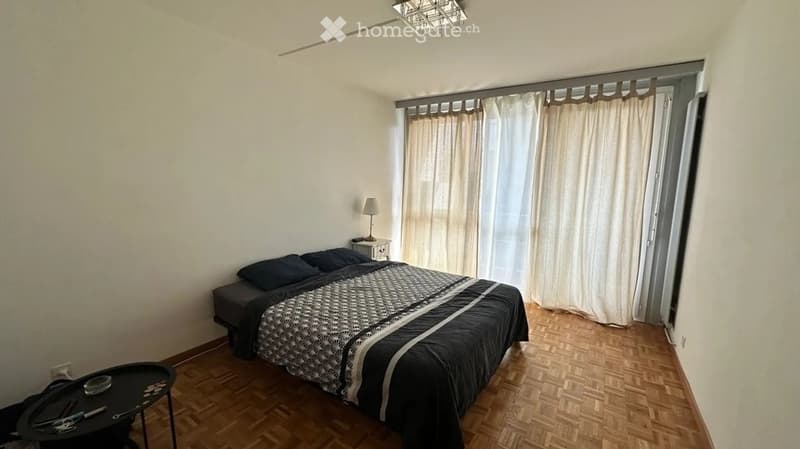 A furnished bedroom with a double bed, nightstand, lamp, and a small table near the window with curtains.