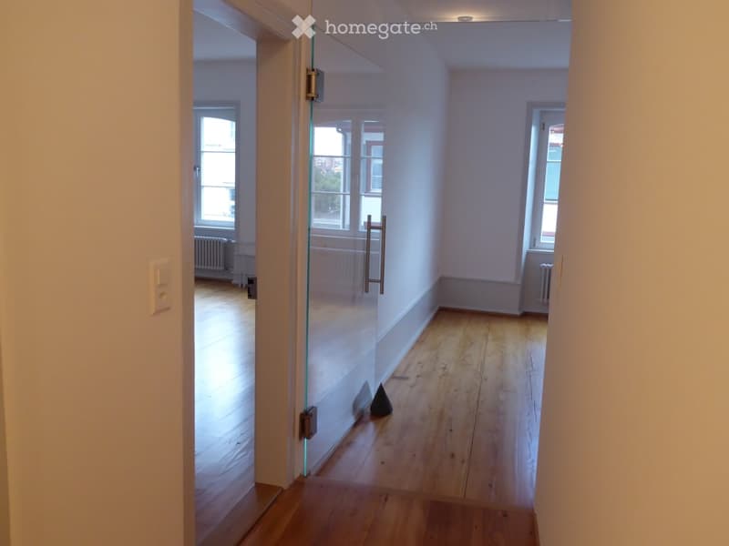 Empty hallway with wooden flooring, glass partition, and visible radiators in adjacent rooms.