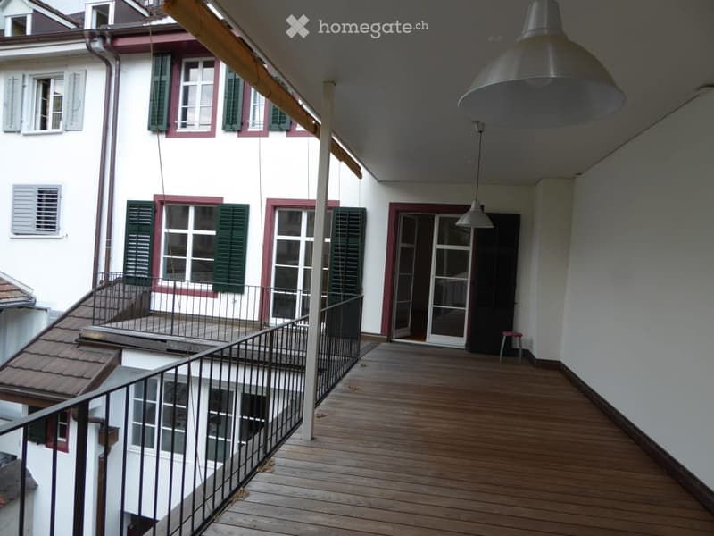 Wooden balcony with metal railing, overlooking a building with green shutters and white walls.