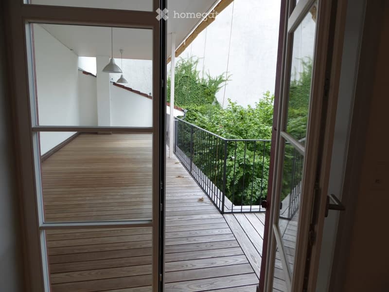 Balcony with wooden decking, glass doors, metal railing, and greenery visible beyond.