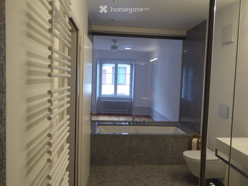 Modern bathroom with a bathtub, toilet, sink, and radiator visible through a glass partition. The space features white walls, a tiled floor, and a window allowing natural light.