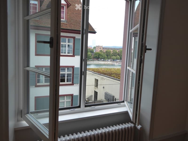 Open window view of a cityscape with buildings, trees, and a body of water in the background. The room has a radiator below the window.
