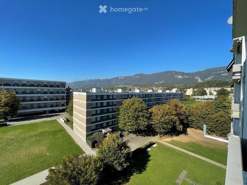 A view from a balcony showing a green lawn, trees, and several apartment buildings in the background with mountains visible in the distance.