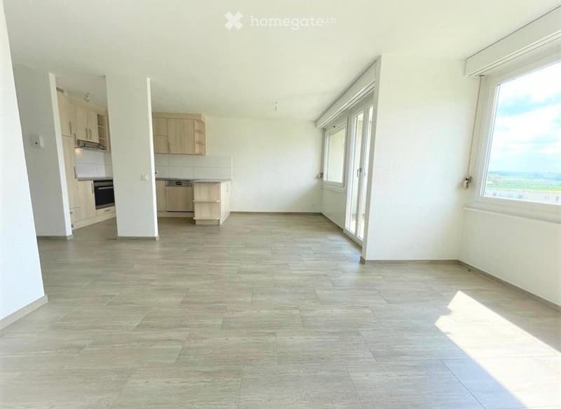 Empty room with tiled floor, white walls, large windows, and an open doorway leading to a kitchen area with cabinets and appliances.