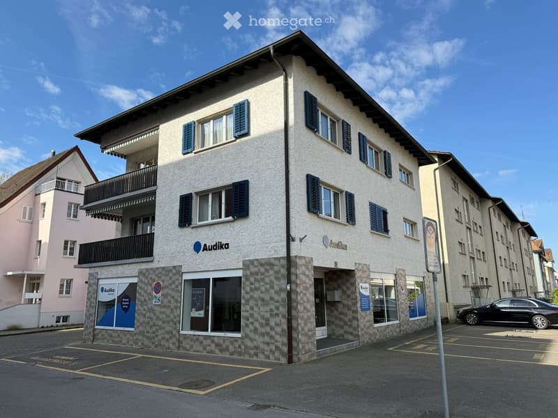 Three-story building with a storefront labeled "Audika" and a balcony on the second floor. The building has multiple windows with shutters and a parking area in front.
