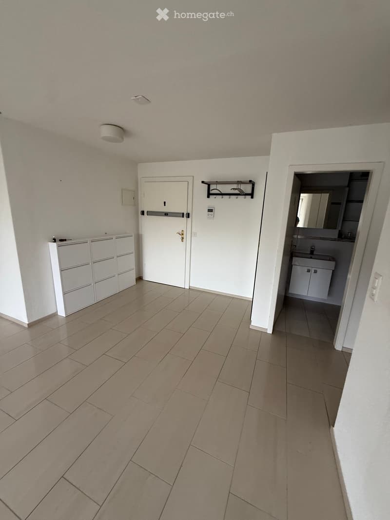 Empty hallway with tiled floor, white walls, a closed door, a coat rack, and an open doorway leading to a small bathroom with a sink and cabinet.