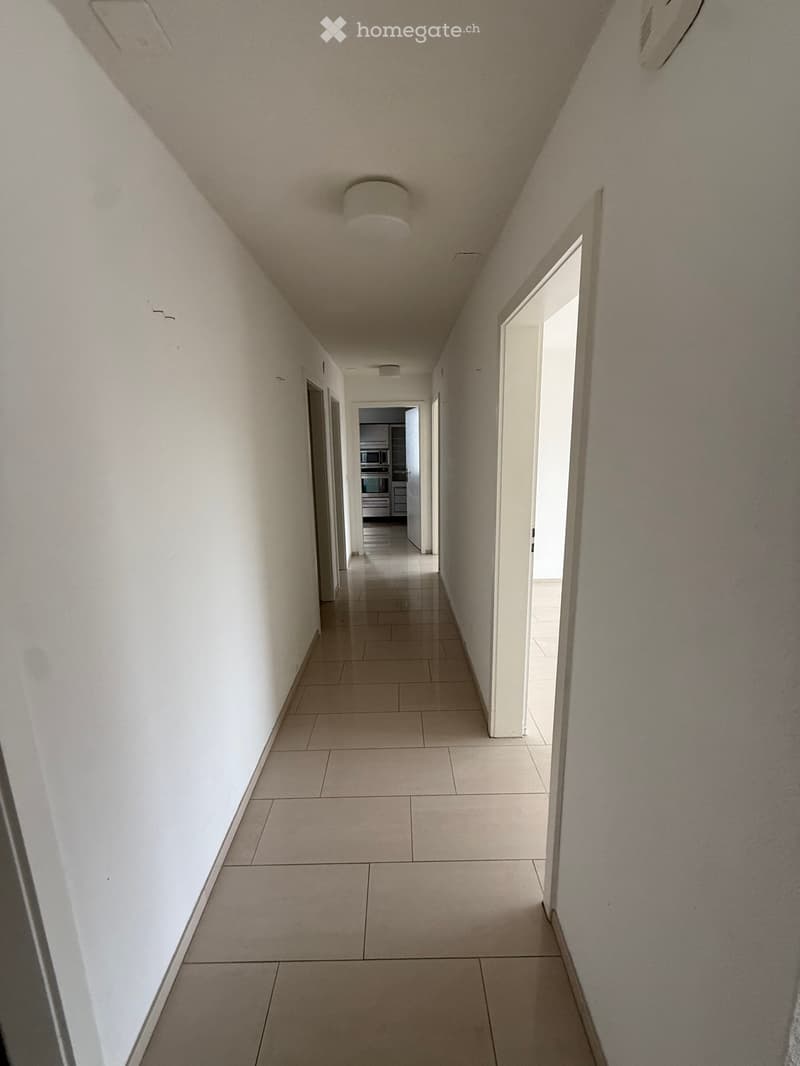 A long, narrow hallway with tiled flooring and white walls, featuring several doors and a visible kitchen area at the end.