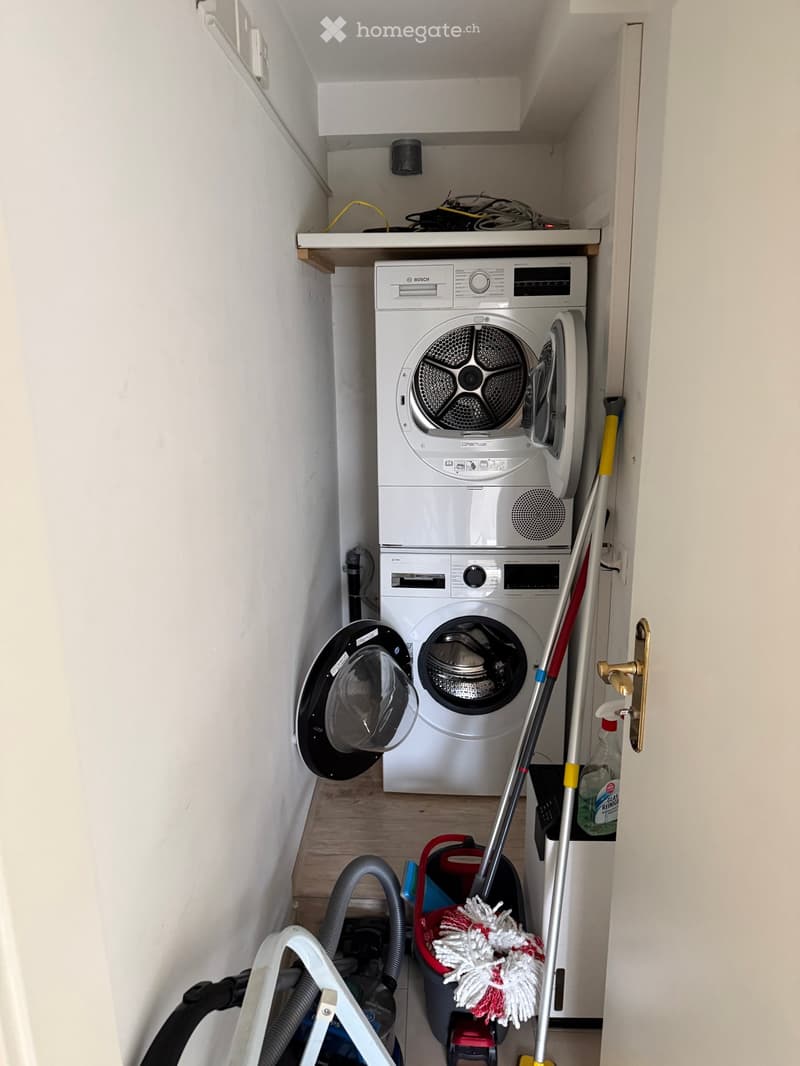 A compact utility room with a stacked washing machine and dishwasher, a mop, vacuum cleaner, and cleaning supplies.