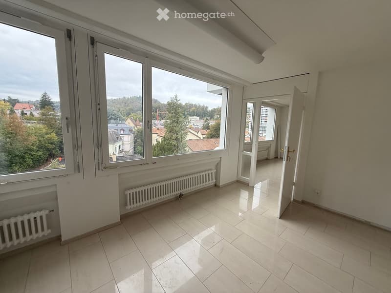 Empty room with tiled floor, large windows offering a view of trees and buildings, radiators along the walls, and an open doorway leading to another space.