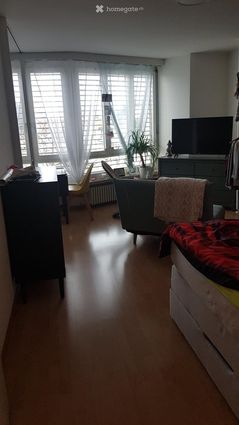 A well-lit living room with wooden flooring, a sofa with a red blanket, a yellow chair, a small table, a television on a cabinet, and a potted plant near the window.