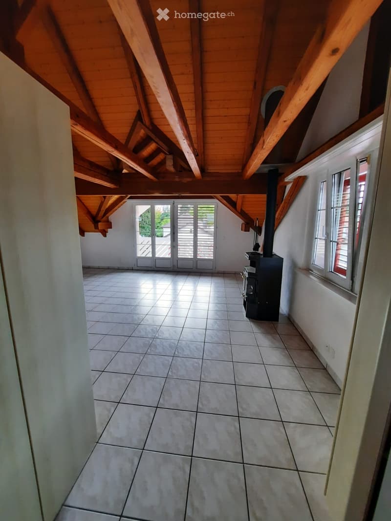 Empty attic space with exposed wooden beams, tiled floor, large windows, and a wood-burning stove.