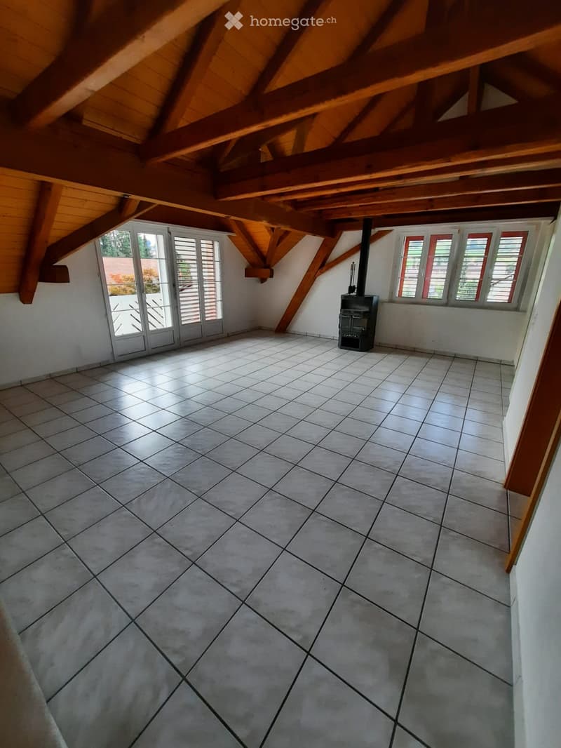 Empty attic space with exposed wooden beams, tiled floor, and a wood-burning stove. Large windows with shutters provide natural light.