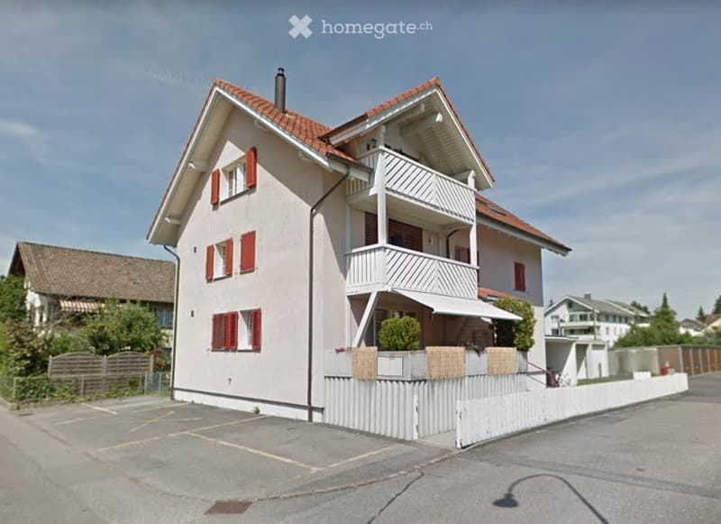 Two-story house with red shutters, white balcony railings, and a parking area.