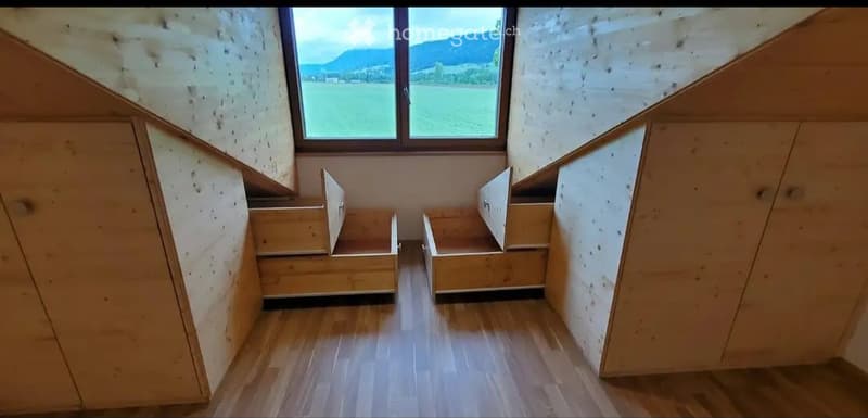 Empty room with wooden flooring, built-in wooden benches along the walls, and a large window offering a scenic view of a field and mountains.