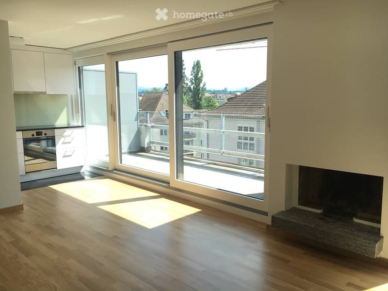 Bright, modern living space with large sliding glass doors opening to a balcony, wooden flooring, fireplace, and a small kitchen area with cabinets and an oven visible.