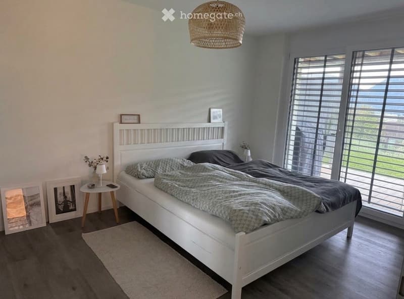 Modern bedroom with a white bed, nightstand, table lamp, and large windows with blinds. The room features a wicker pendant light and a small rug on the floor.