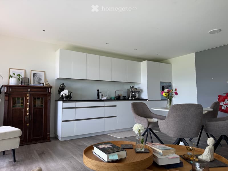 Modern open-plan living and dining area with a grey sofa, wooden coffee table, dining chairs, and a kitchen with white cabinets and stainless steel appliances.