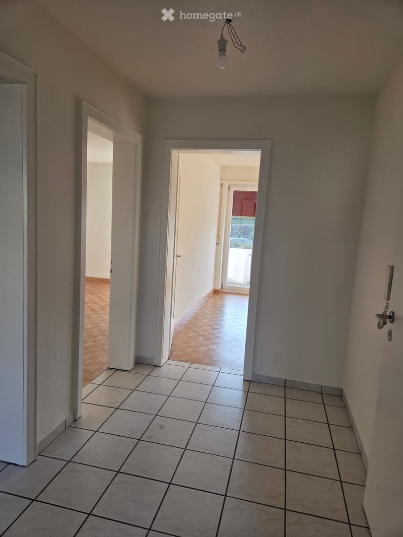 Empty hallway with tiled floor and wooden flooring visible in adjacent rooms. Natural light from an open door at the end.