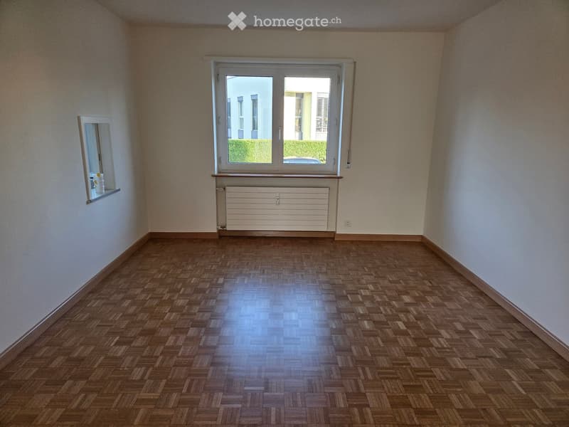 Empty room with wooden parquet flooring, white walls, large window with white frame, radiator below window, and small shelf with a few items on the left side.