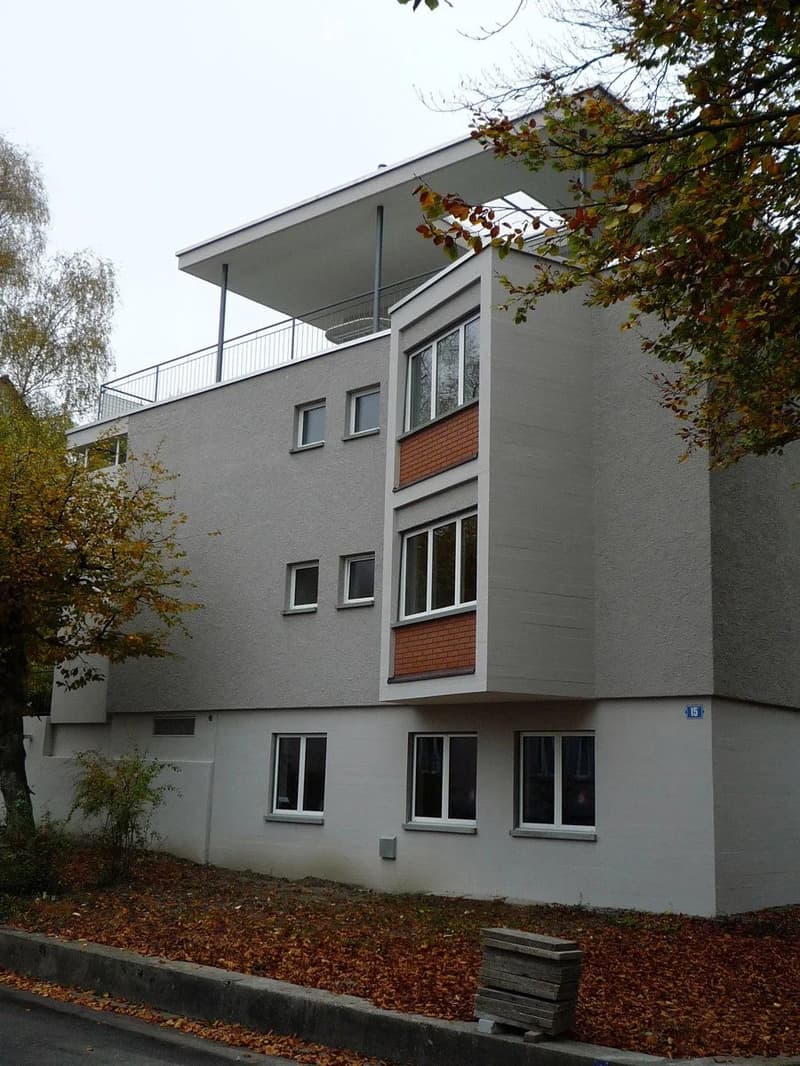Modern two-story building with grey facade, large windows, and a flat roof with a small balcony area.