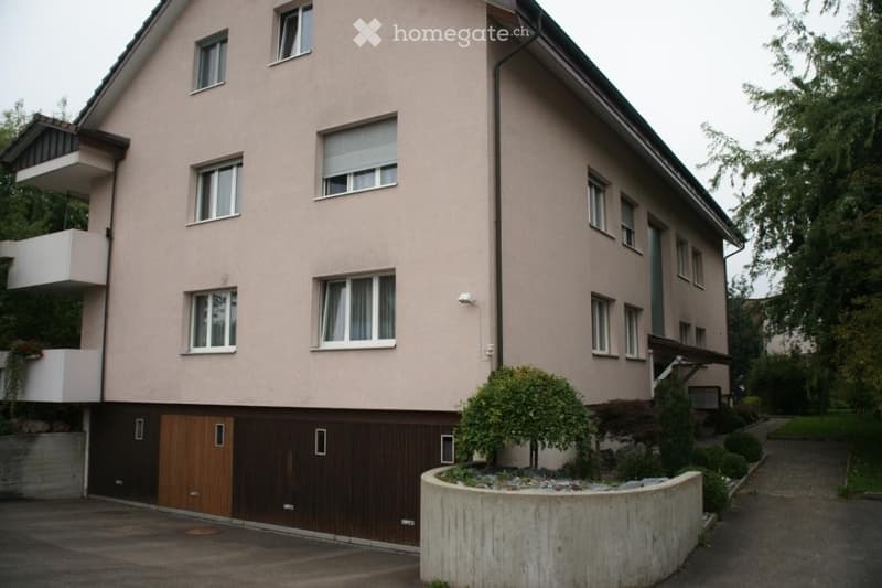 Two-story residential building with beige facade, multiple windows, and a balcony on the upper floor. Features a wooden garage door and small landscaped area in front.