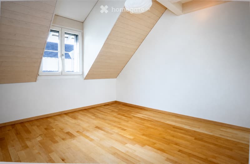 Empty room with wooden flooring, white walls, and a slanted wooden ceiling. A single window with white frame is visible on one wall.