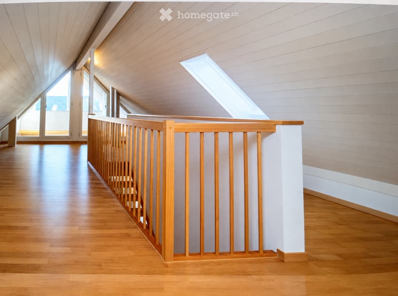 Bright, modern hallway with wooden flooring, white walls, and a wooden railing leading to a staircase. Large windows allow natural light to fill the space.