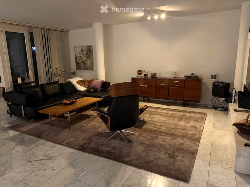 Modern living room with a black leather sofa, wooden coffee table, leather armchair, and a large cabinet. The room features a marble tile floor, a large area rug, and decorative items including a candle holder and a fruit bowl.