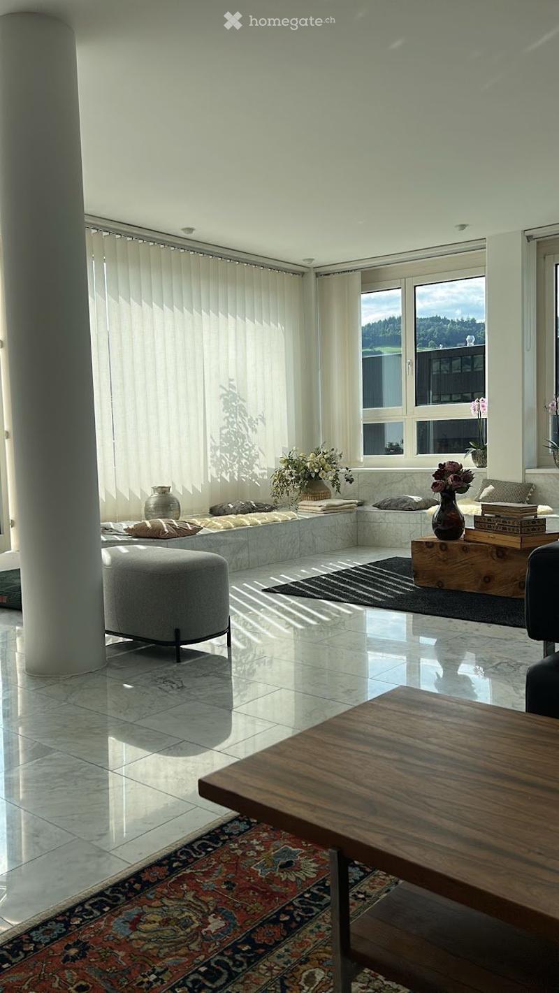 Bright living room with large windows, white walls, polished tile floor, and a mix of modern and traditional furniture including a sofa, coffee table, ottoman, and decorative vases.