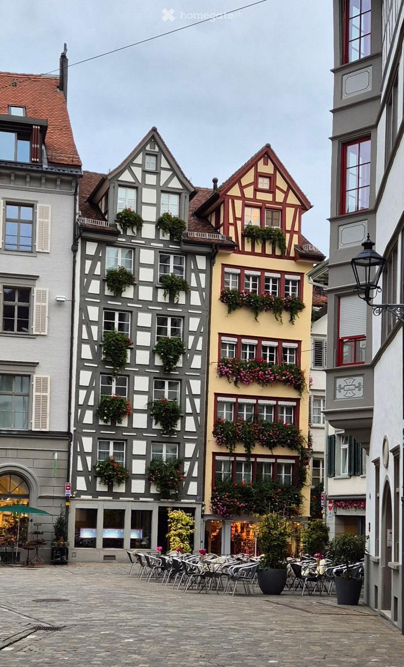 A charming European town square with traditional half-timbered houses, flower boxes, outdoor seating, and cobblestone pavement.