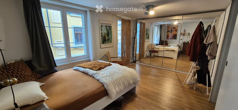 Bright bedroom with wooden flooring, large window with view, bed with pillows and blanket, mirror, chair, and artwork on wall.