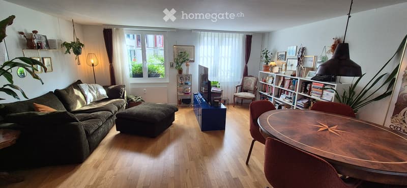 A cozy living room with a dark green sectional sofa, ottoman, wooden dining table with chairs, bookshelf, and decorative plants.